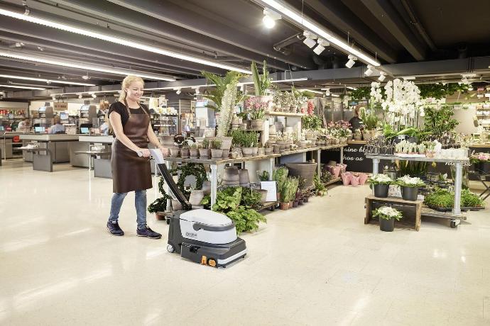 Compact floor scrubber cleaning a retail showroom with display plants and shelving